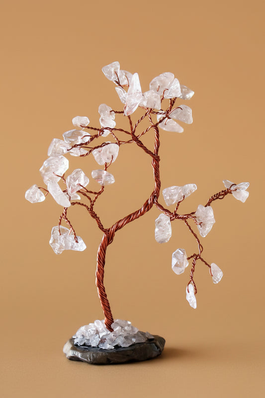 A crystal tree decorative sculpture with clear quartz-like stones on copper-colored wire branches, displayed on a small dark base against a warm tan background.