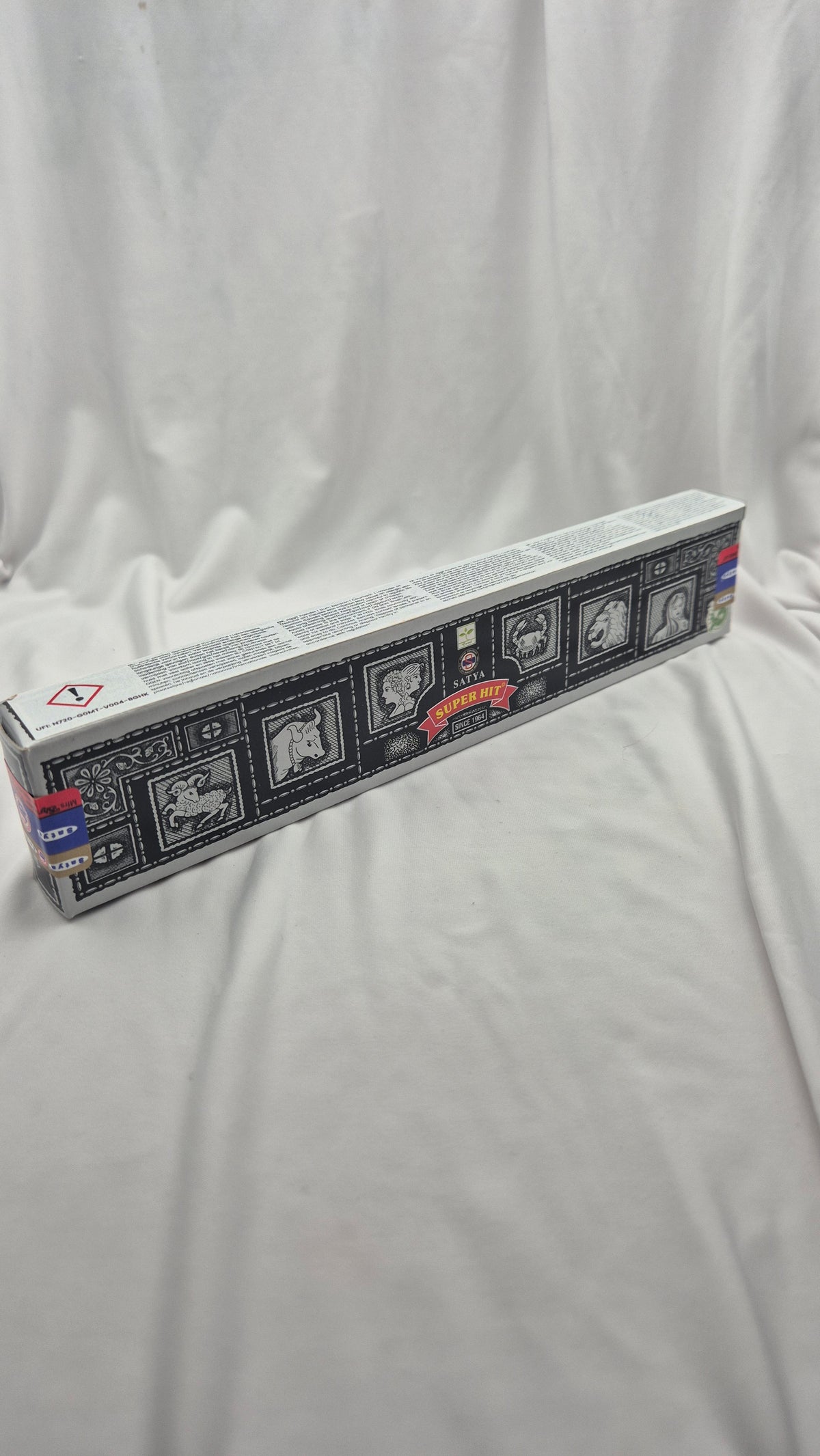 A long rectangular product box with a black, white, and gray illustrated design and small red accents, photographed on a white fabric backdrop.