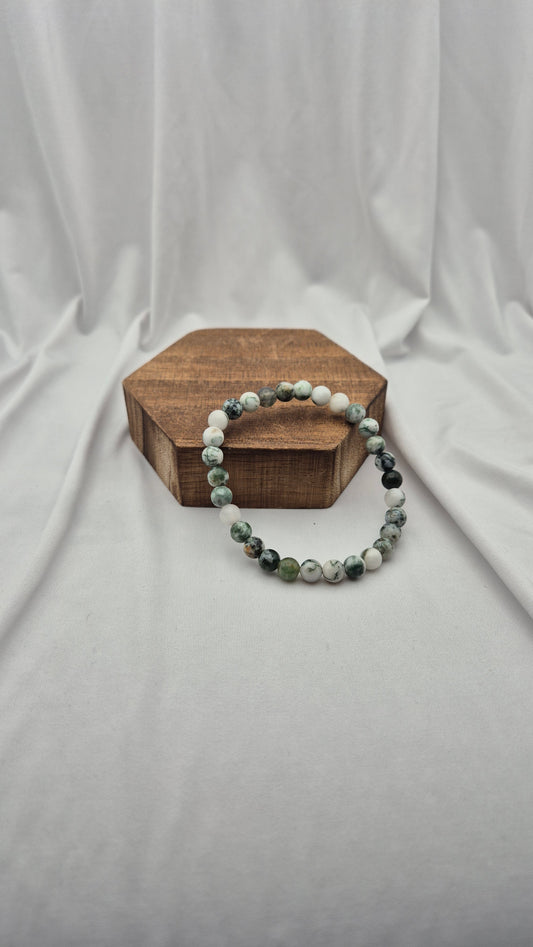 A beaded stretch bracelet made of green, white, and dark gray marbled beads, displayed on a small hexagonal wooden block against a draped white fabric backdrop.