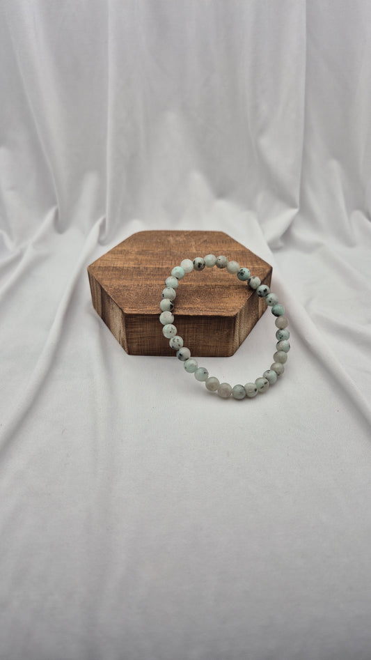 A beaded stretch bracelet made of light gray-green marbled beads, draped over a hexagonal wooden block on a draped white fabric background.