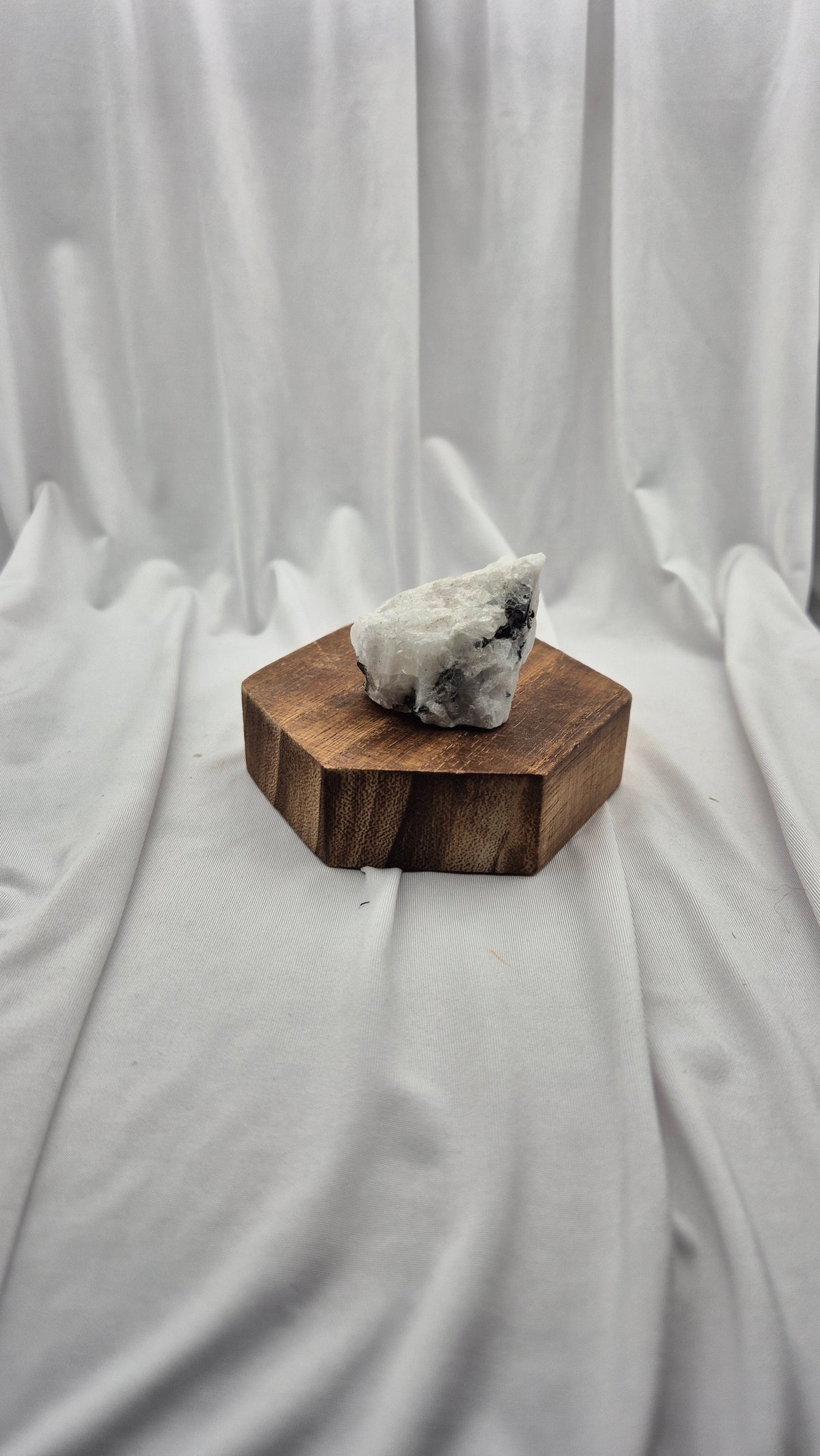 A raw white crystal cluster with dark inclusions sits on a hexagonal dark brown wooden block, set against a draped white fabric backdrop.