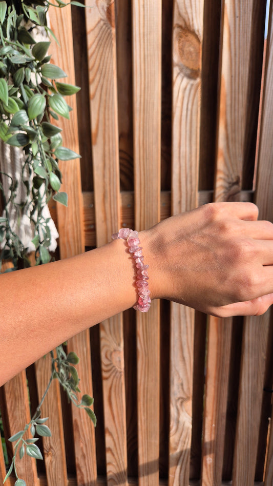 A pink gemstone chunk bracelet worn on a person's wrist, shown against a wooden slat wall with green trailing leaves on the left.