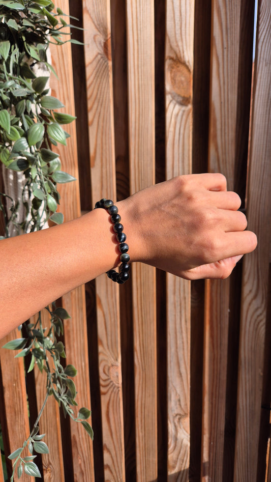 A person's hand and forearm wearing a black beaded bracelet, posed in front of a vertical wooden slat fence with green trailing leaves on the left.