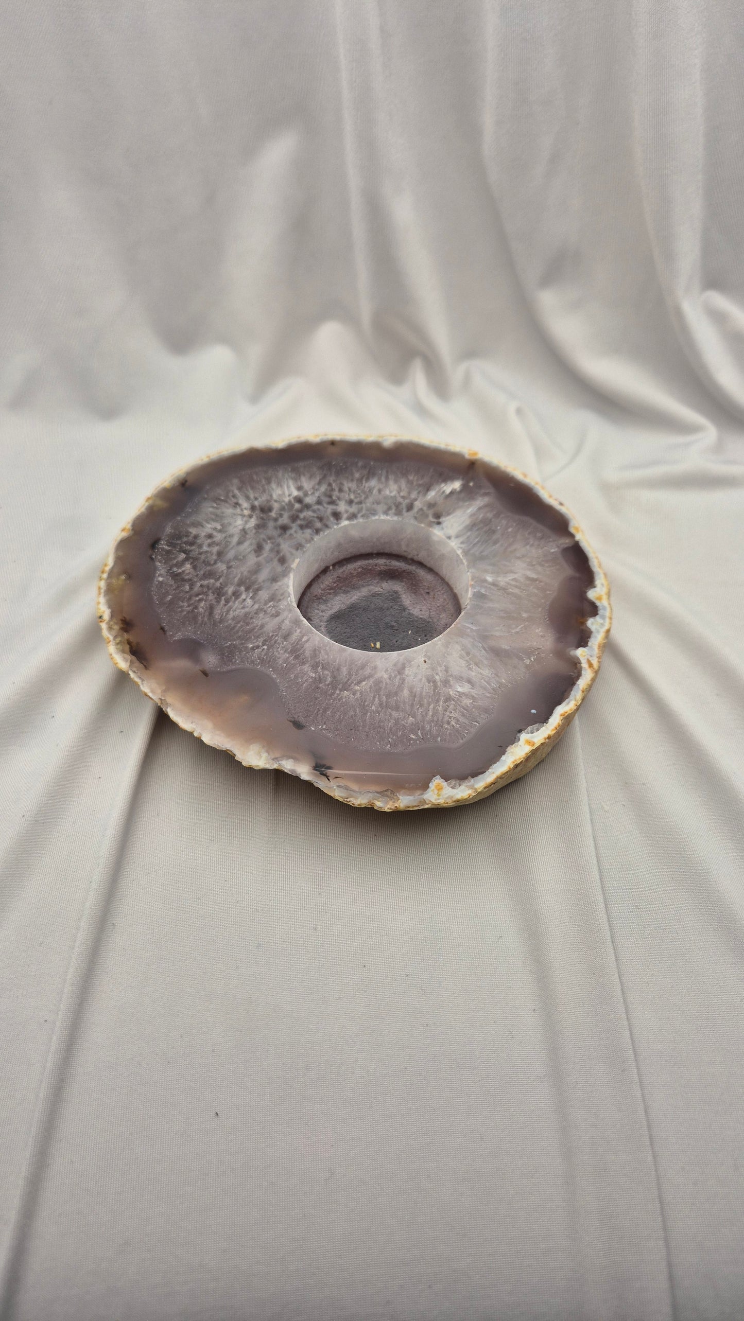 A circular agate stone bowl with a natural geode center, featuring concentric bands of gray, purple, and brown, displayed on draped white fabric.