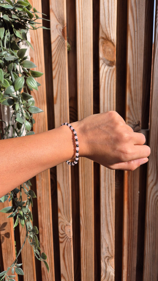 A beaded bracelet with small dark beads and white accents worn on a person's wrist, shown against a wooden slat wall with green foliage on the left.