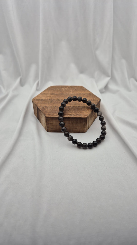 A dark beaded bracelet arranged in a loop around a hexagonal wooden display block, set against a draped white fabric backdrop.