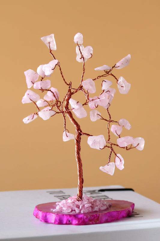 A pink crystal tree of life decorative sculpture made of copper wire and light pink quartz stones, displayed on a round magenta agate base against a warm tan background.