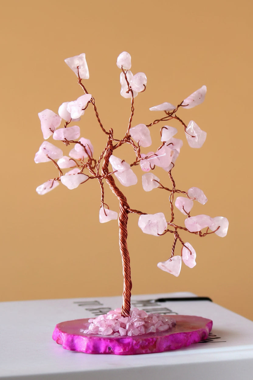 A pink crystal tree of life decorative sculpture made of copper wire and light pink quartz stones, displayed on a round magenta agate base against a warm tan background.
