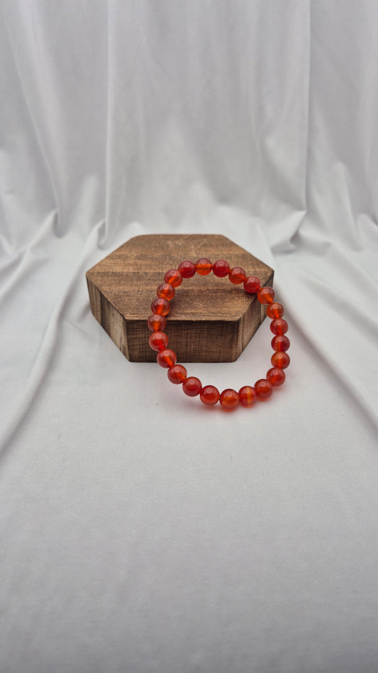 A beaded stretch bracelet made of translucent orange-red round beads, displayed on a small hexagonal wooden block against a draped white fabric backdrop.