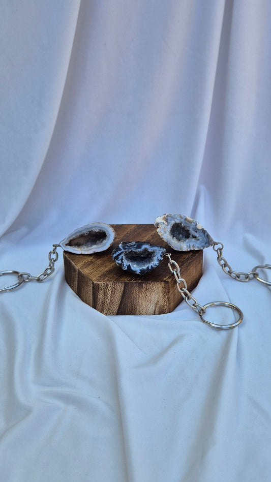 Three silver chain keychains with natural agate geode pendants rest on a small wooden block against a draped white fabric backdrop.