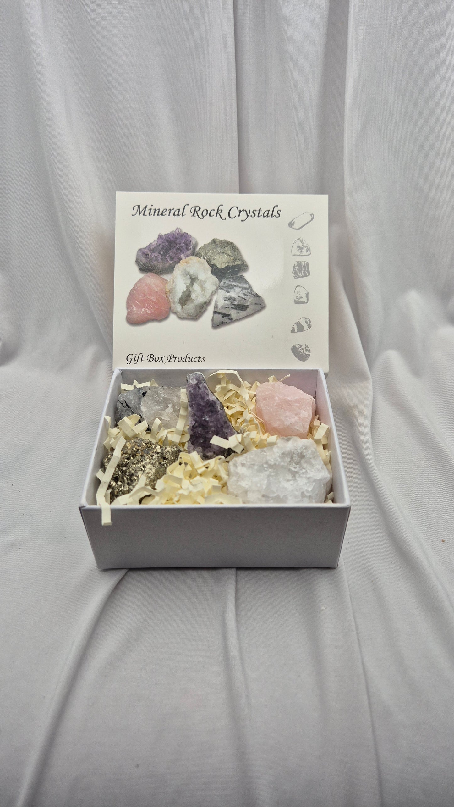 A gift box of mineral and crystal rocks displayed on a draped white fabric background. The open white box contains several raw crystals and rocks nestled in shredded paper filler, with the lid printed with 'Mineral Rock Crystals' and small illustrations.