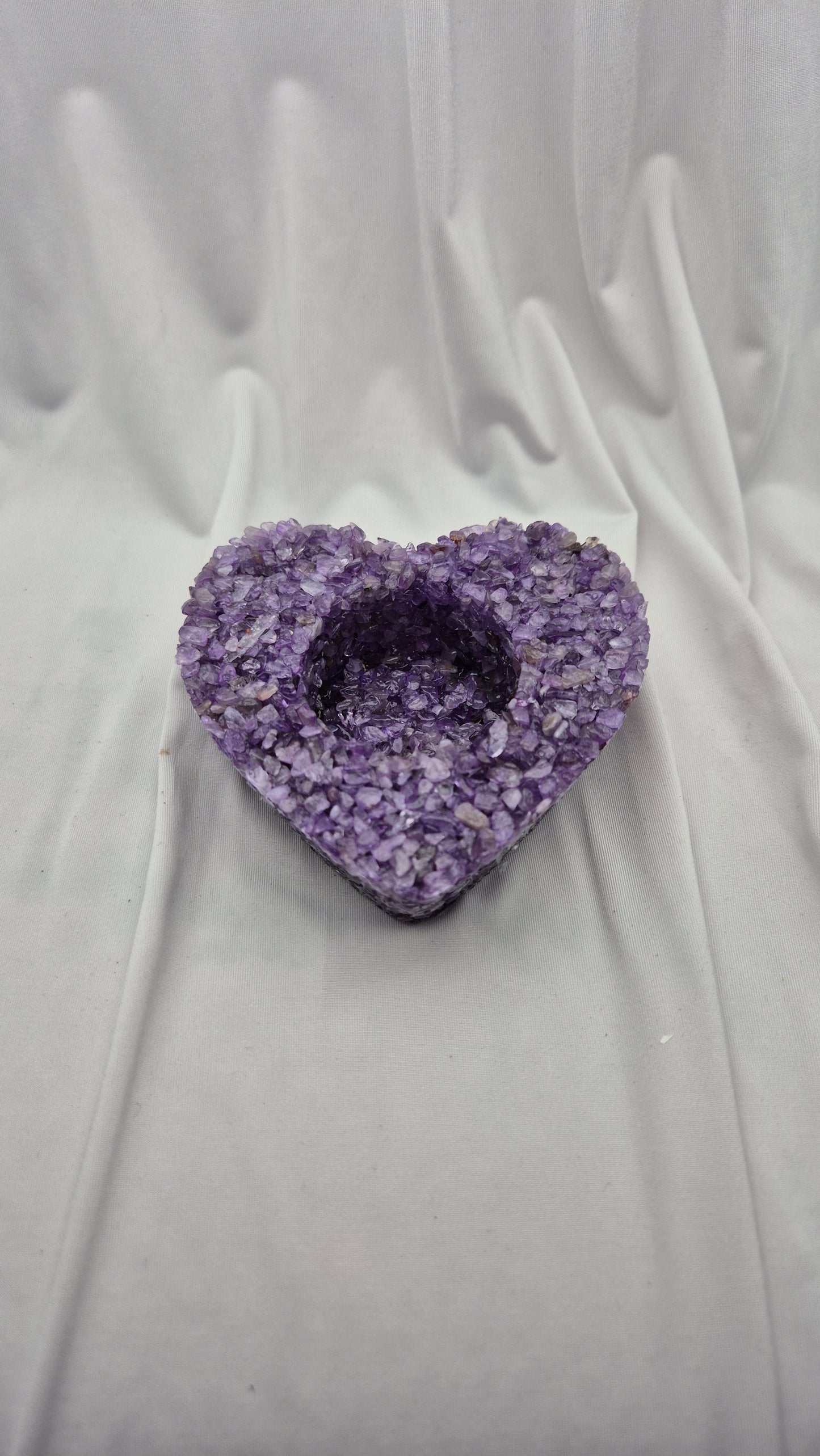 A heart-shaped purple crystal geode candle holder with a textured, glittering amethyst exterior and a hollow center, displayed on a draped white fabric background.