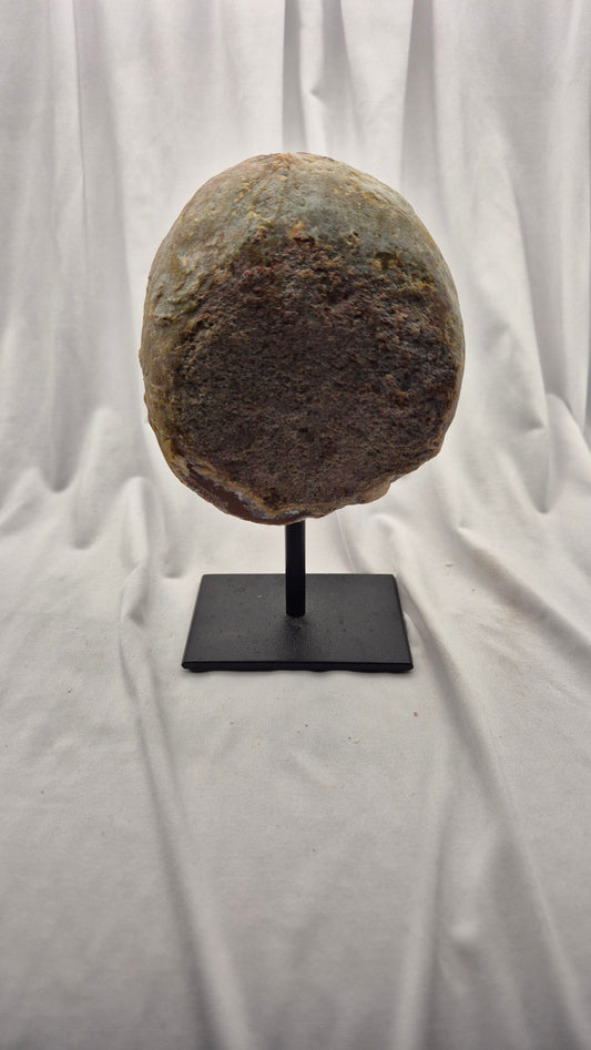 A large, oval-shaped natural stone or rock specimen displayed on a black metal stand against a draped white fabric backdrop.