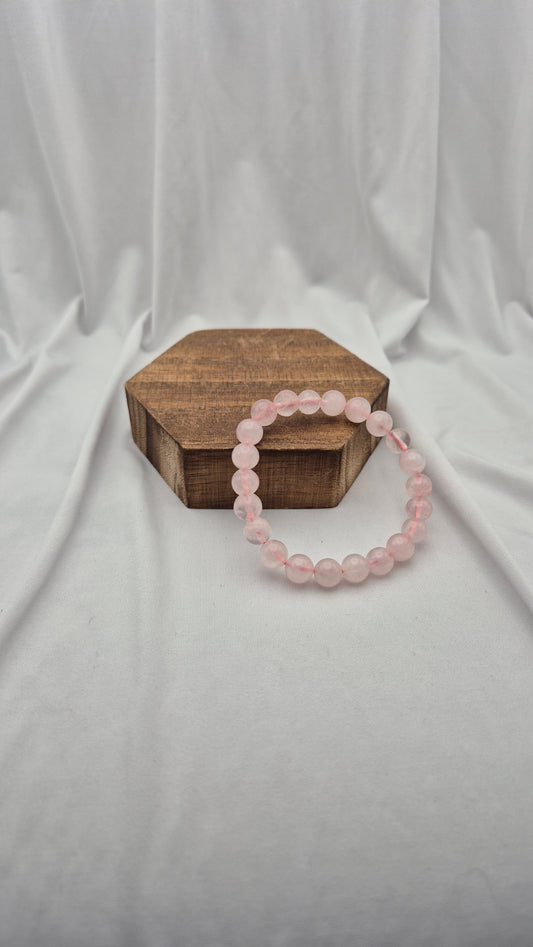 A pink beaded bracelet made of smooth, round stones, displayed on a small hexagonal wooden box against a draped white fabric background.