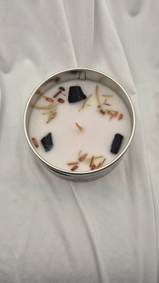 A round metal tin candle filled with white wax and topped with dried botanical pieces and black stones, set against a draped white fabric background.