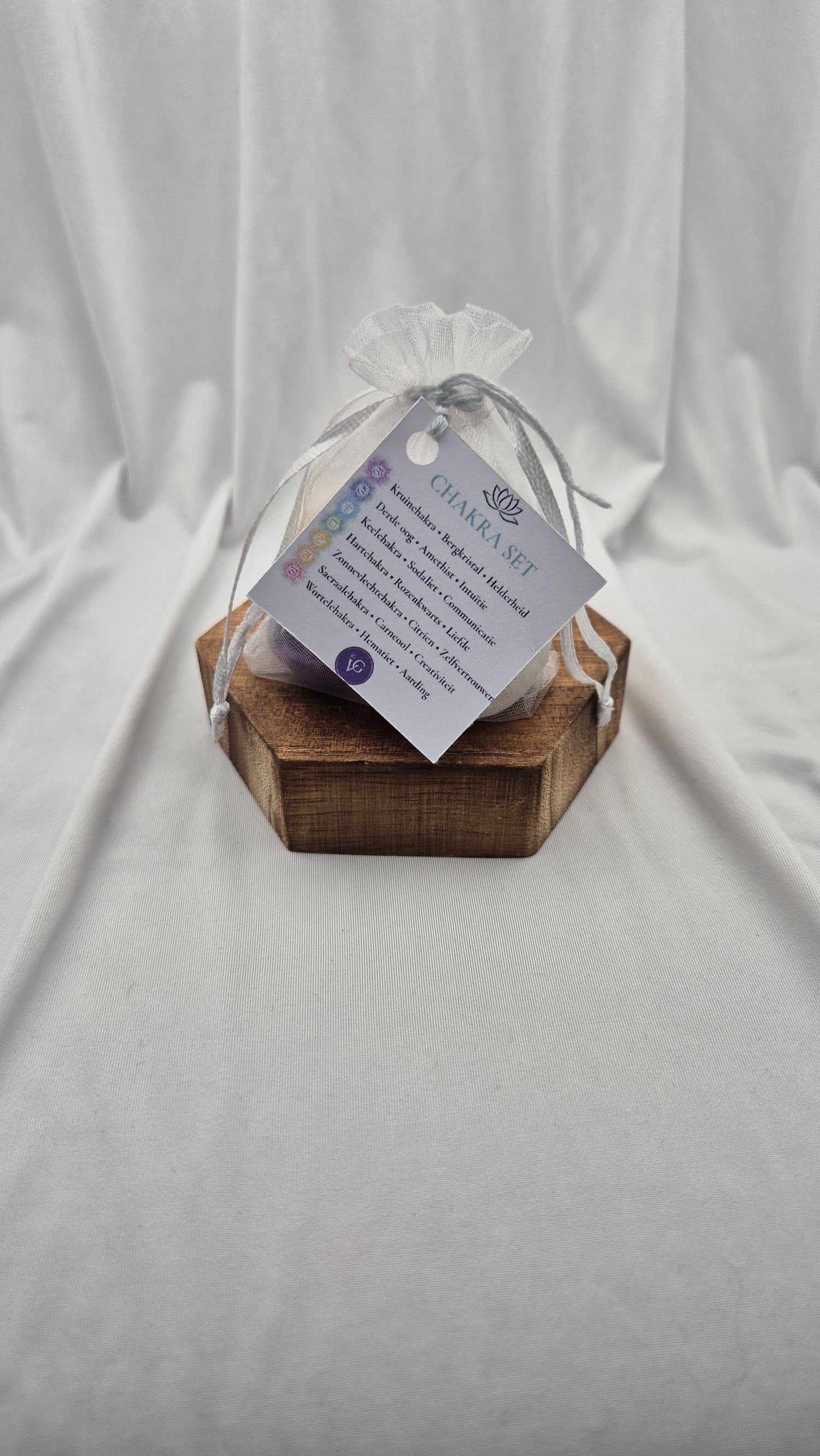 A small clear mesh gift bag tied with a sheer ribbon, resting on a hexagonal wooden block. A printed tag attached to the bag reads 'CHANEL SET' with additional text below, photographed against draped white fabric.