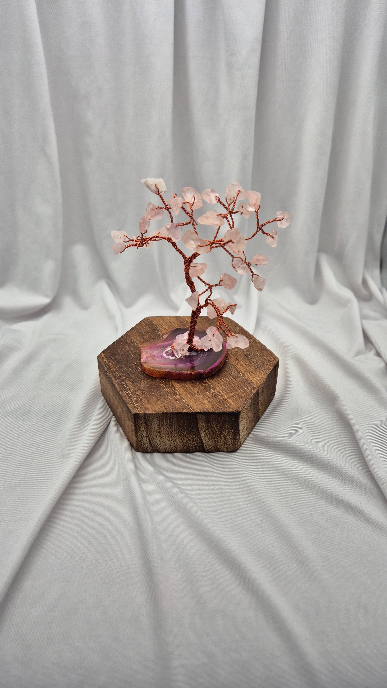 A cherry blossom crystal tree miniature with pink rose quartz blossoms and copper-colored branches, displayed on a polished purple agate slice and a hexagonal wooden block against a draped white fabric background.