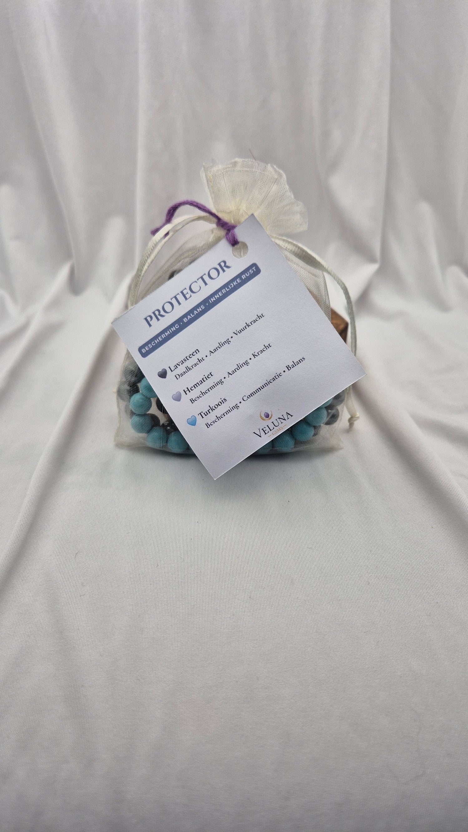A small clear gift bag tied with a purple ribbon, containing a beaded bracelet and a white paper tag that reads 'PROTECTOR' and 'I VOWED TO KEEP YOU SAFE.' The bag is placed on draped light blue fabric.