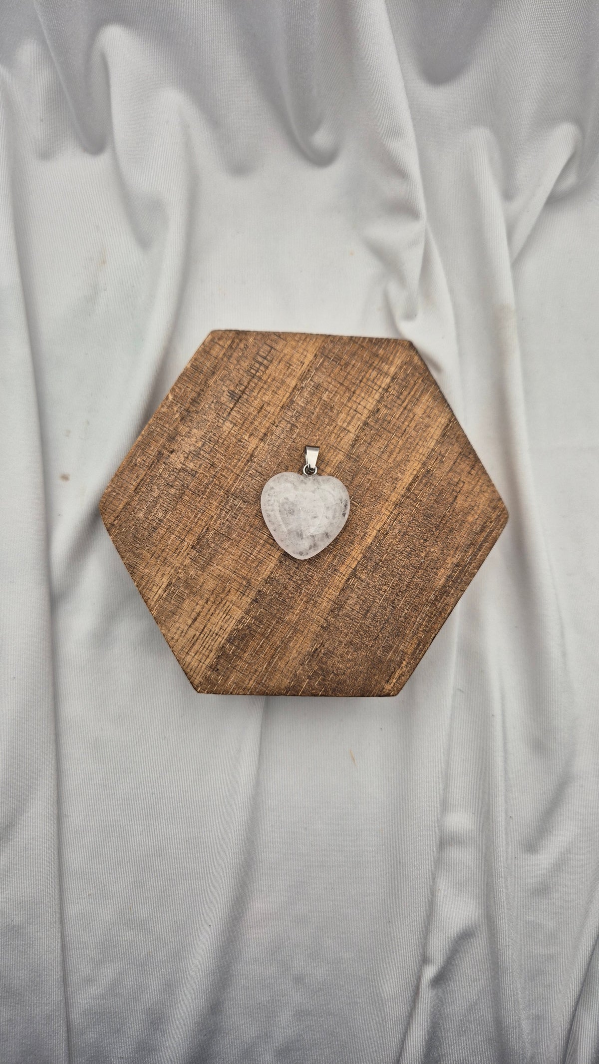 A heart-shaped clear crystal pendant on a silver bail, displayed on a hexagonal wooden block over a draped light gray fabric background.