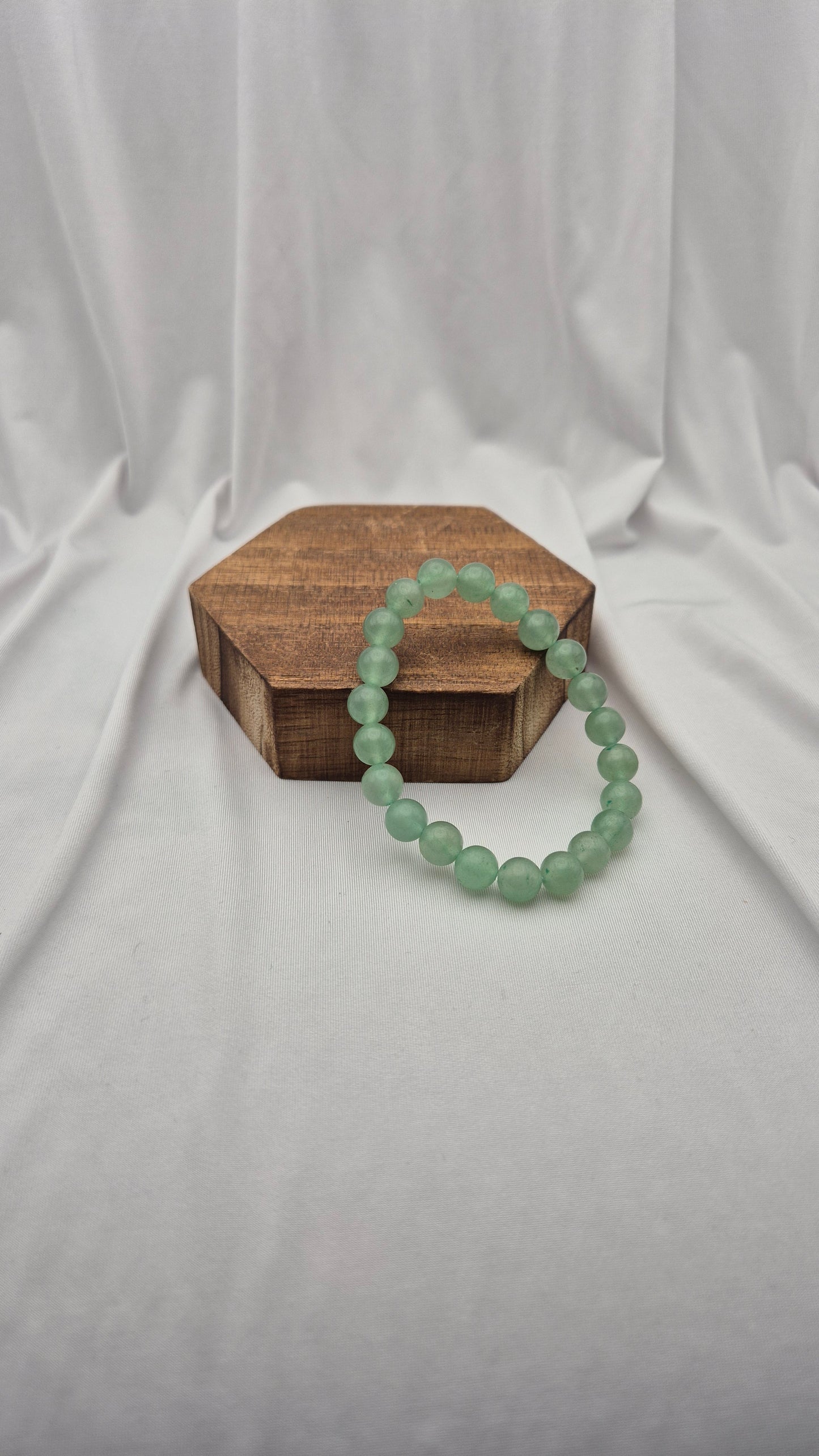 A light green beaded bracelet draped over a small hexagonal wooden box, set against a draped white fabric backdrop.