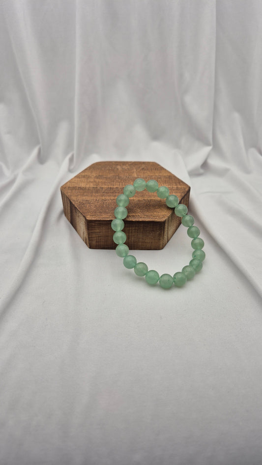 A light green beaded bracelet draped over a small hexagonal wooden box, set against a draped white fabric backdrop.