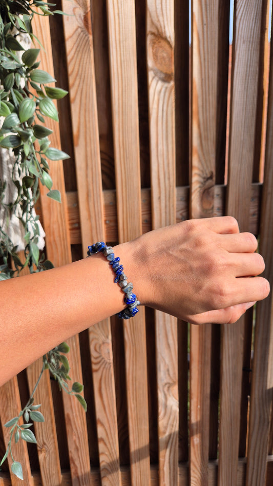 A person's hand and forearm wearing a beaded bracelet with blue and white stones, posed in front of a wooden slat fence with green foliage on the left.