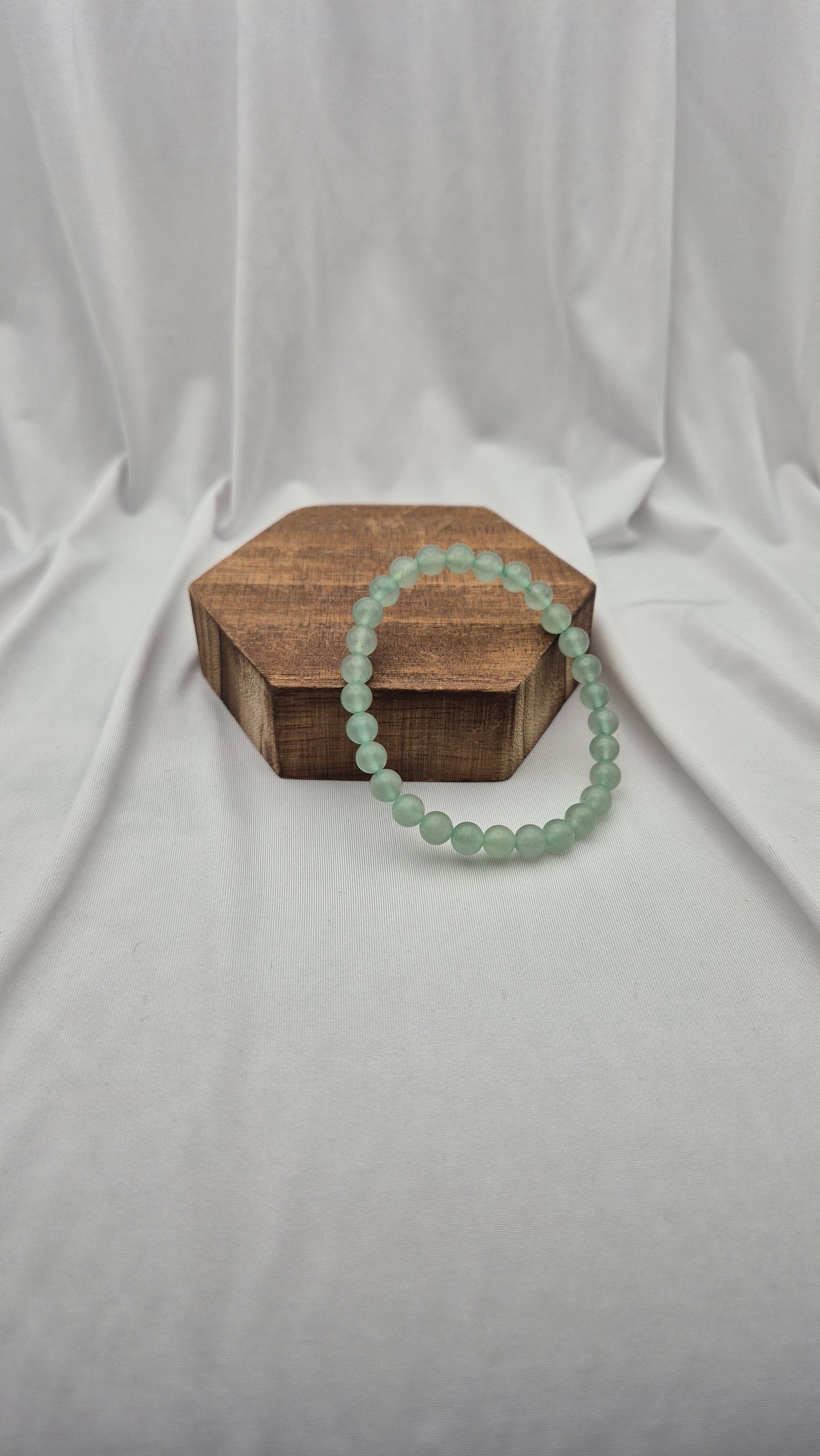 A light green beaded bracelet draped over a small hexagonal wooden box on a draped white fabric background.