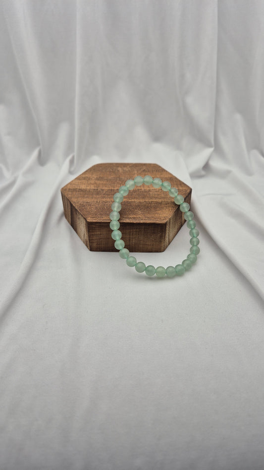A light green beaded bracelet draped over a small hexagonal wooden box on a draped white fabric background.