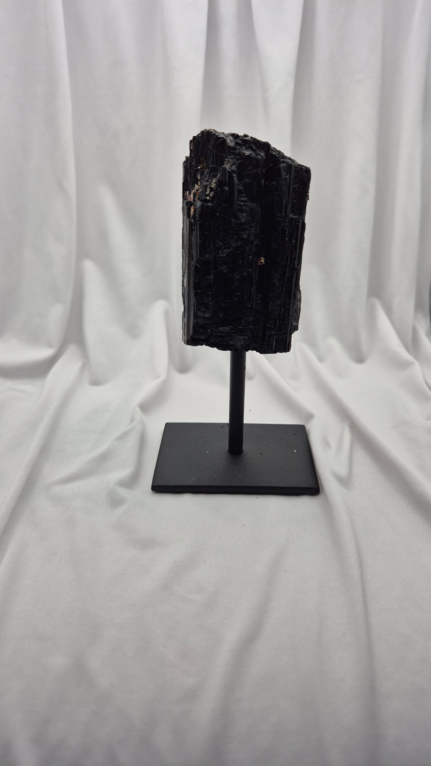 A black mineral specimen or raw gemstone mounted on a black metal display stand, set against a draped white fabric backdrop.