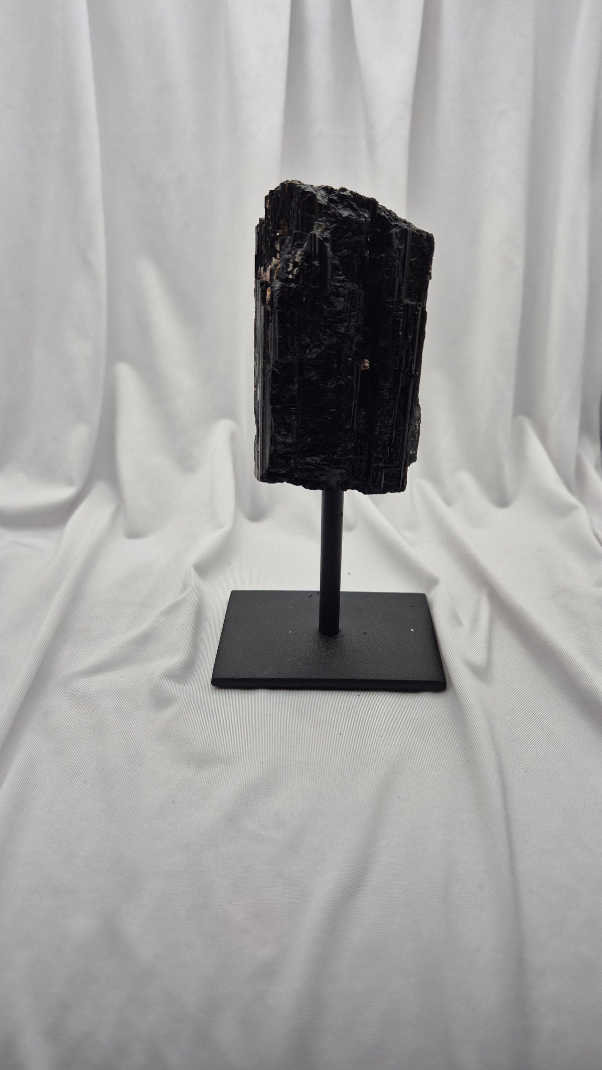 A black mineral specimen or raw gemstone mounted on a black metal display stand, set against a draped white fabric backdrop.