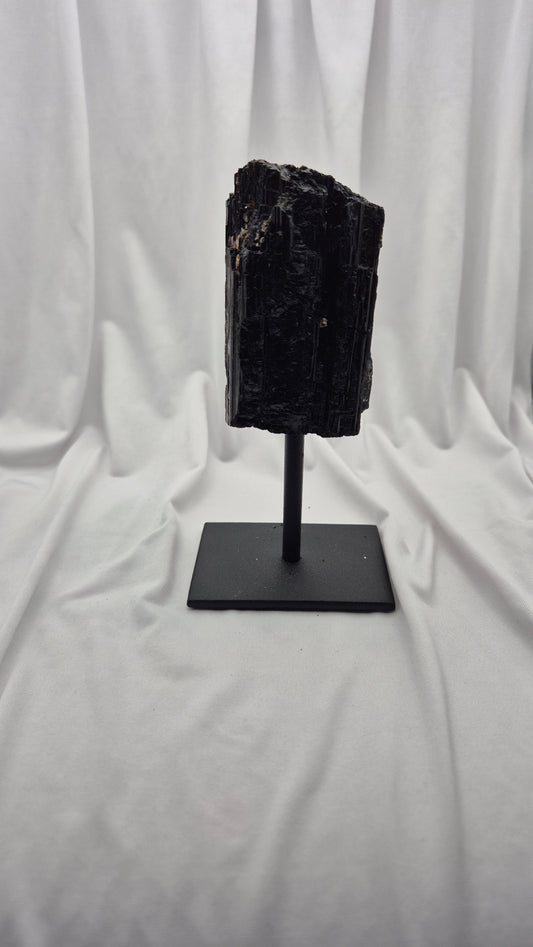 A black mineral specimen or raw gemstone mounted on a black metal display stand, set against a draped white fabric backdrop.