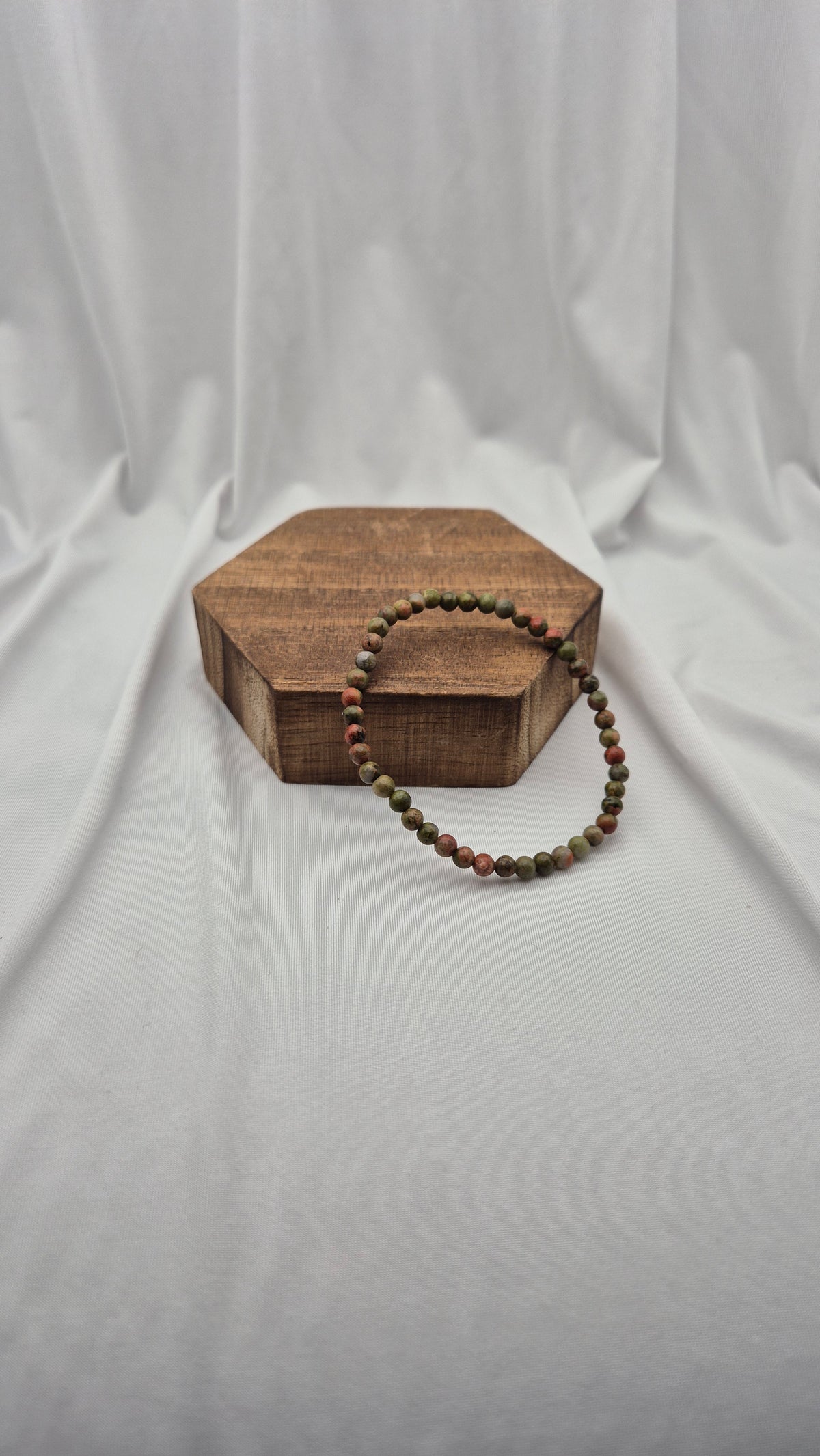 A beaded stretch bracelet with alternating red and green beads, draped over a hexagonal wooden block against a draped white fabric backdrop.