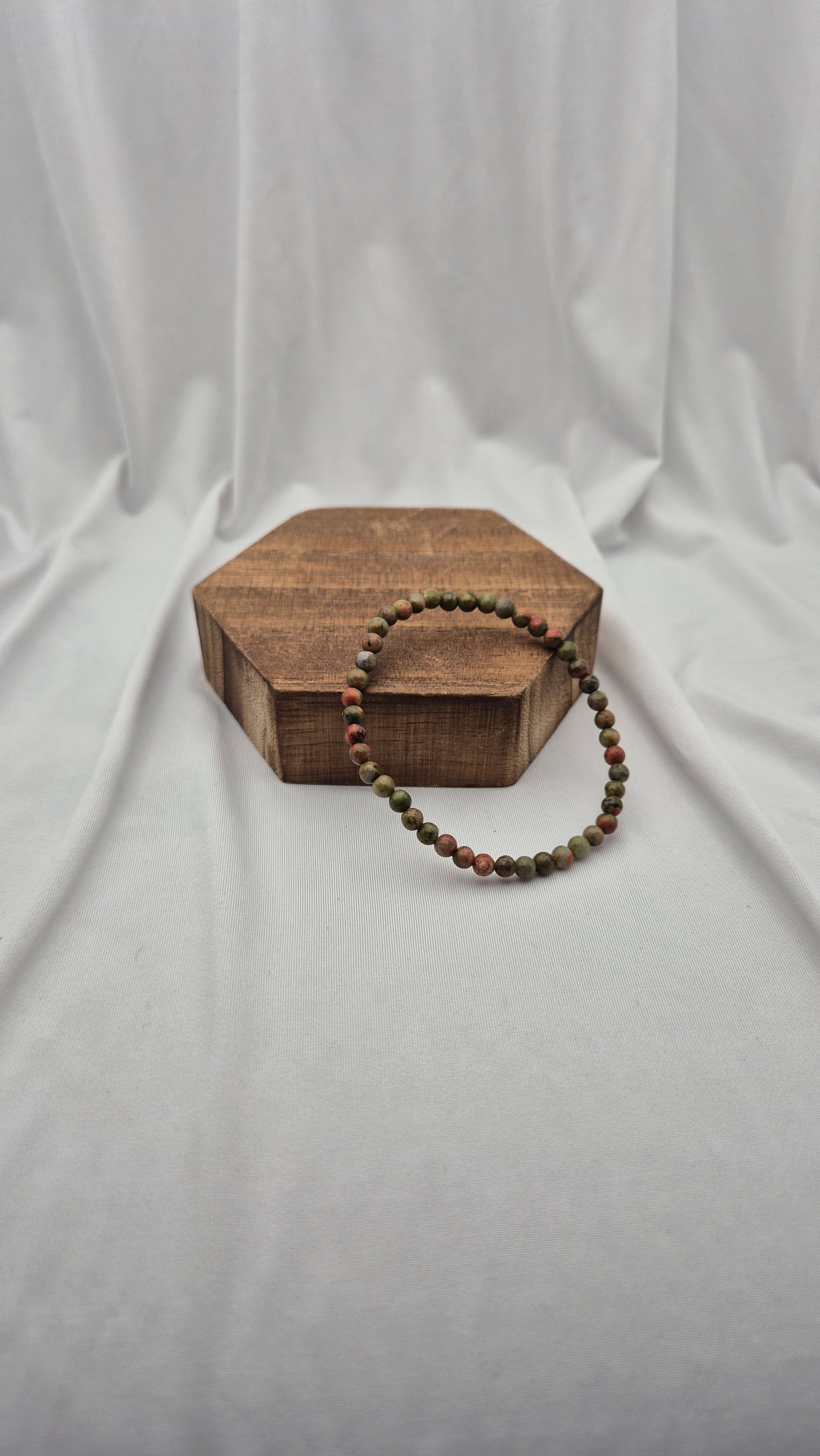 A beaded stretch bracelet with alternating red and green beads, draped over a hexagonal wooden block against a draped white fabric backdrop.
