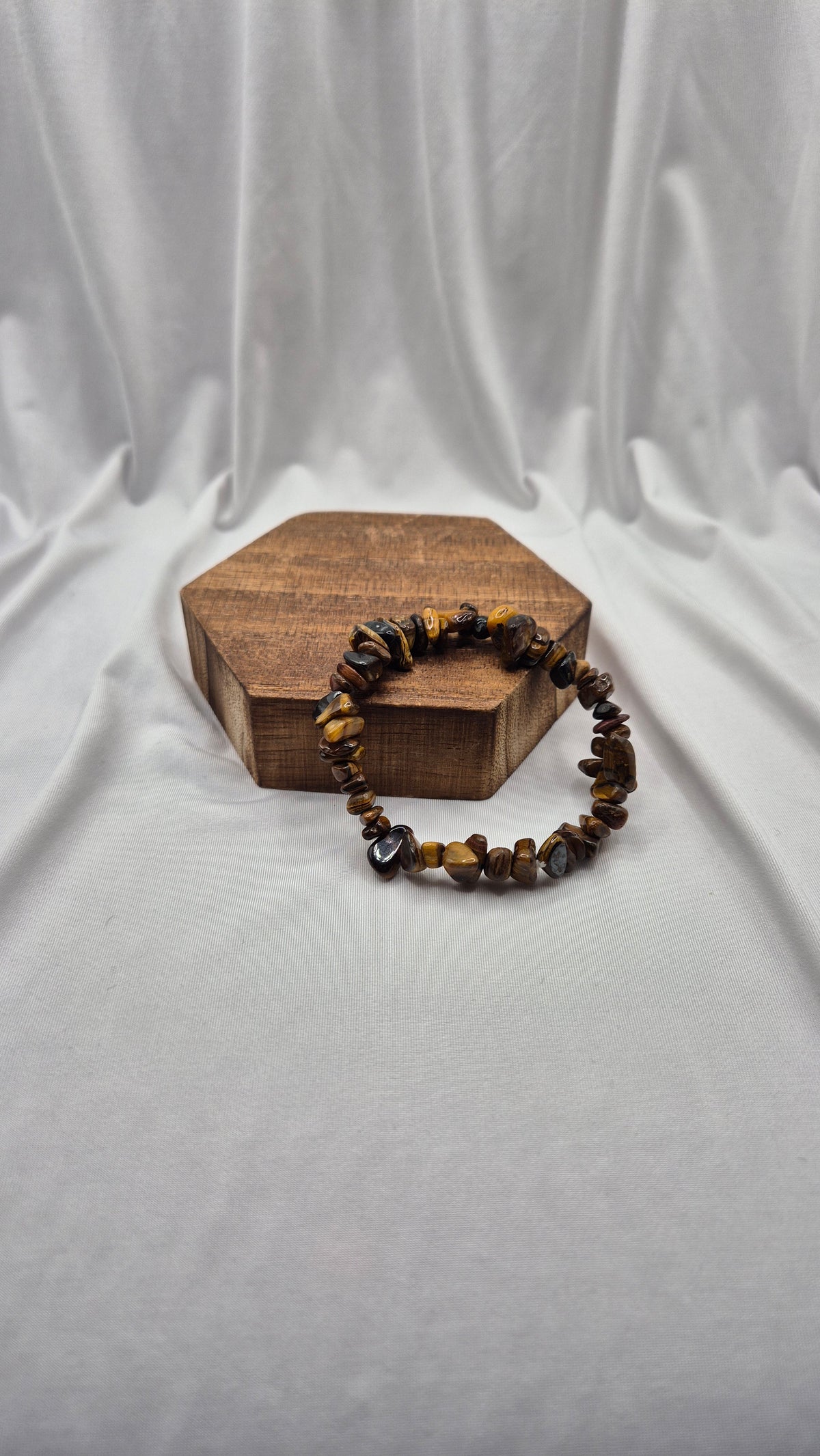 A beaded bracelet made of polished tiger's eye stones in brown and gold tones, resting on a small hexagonal wooden block against a draped white fabric backdrop.