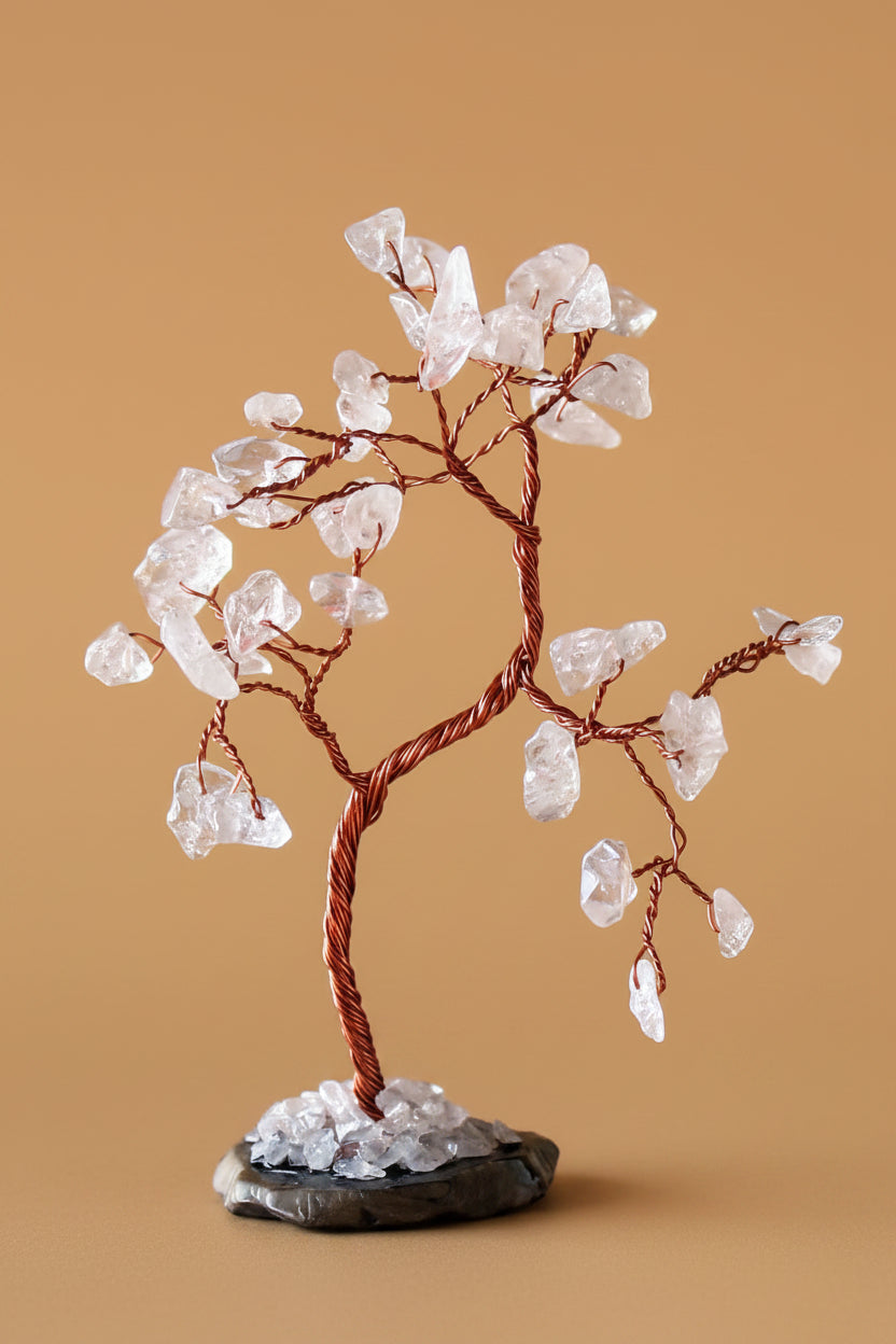 A crystal tree decorative sculpture with clear quartz-like stones on copper-colored wire branches, displayed on a small dark base against a warm tan background.