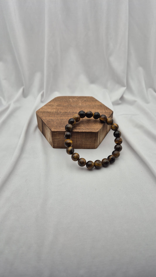 A beaded bracelet made of dark brown and black tiger's eye beads, draped over a small hexagonal wooden block on a draped white fabric background.