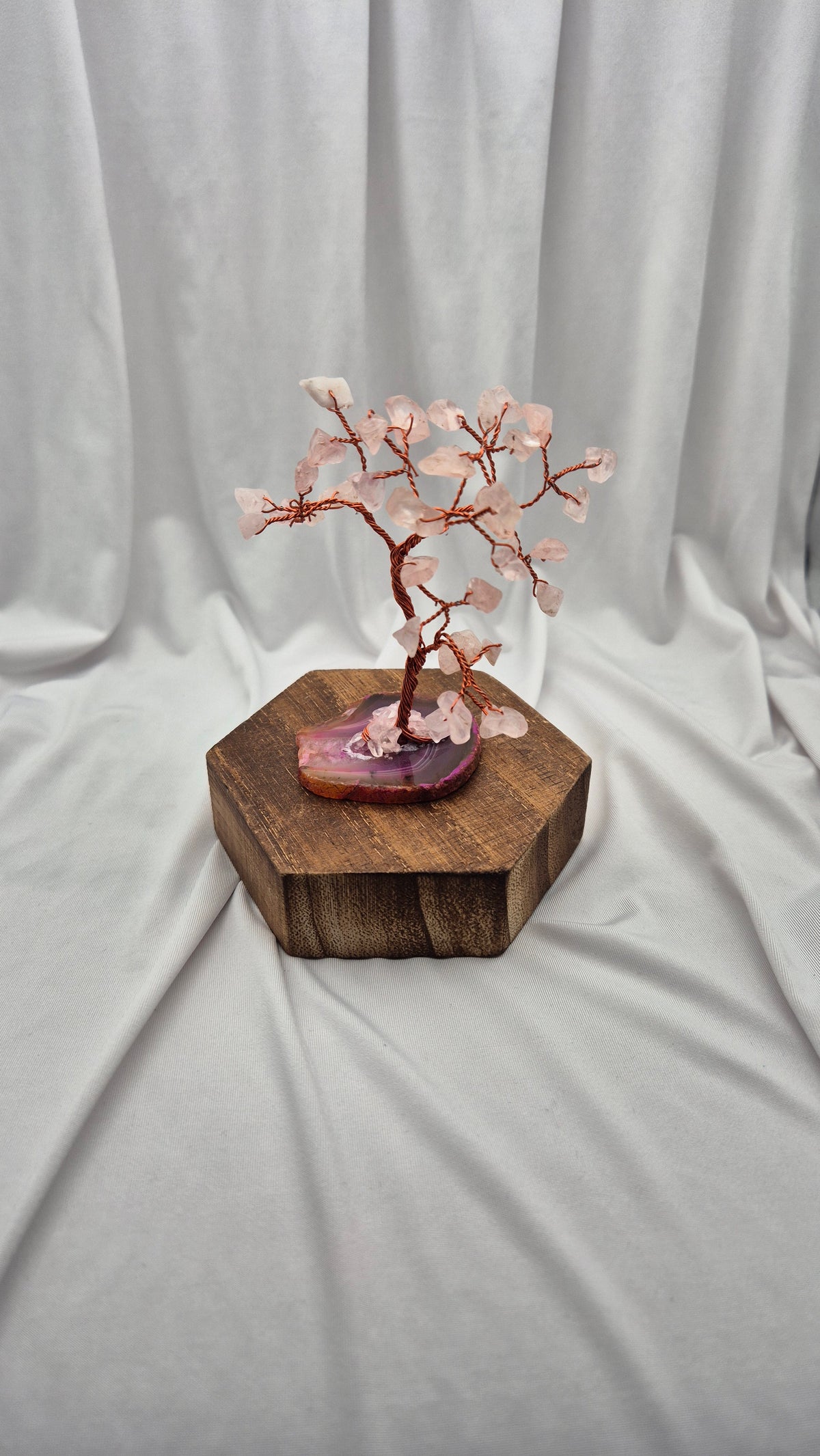A cherry blossom crystal tree miniature with pink rose quartz blossoms and copper-colored branches, displayed on a polished purple agate slice and a hexagonal wooden block against a draped white fabric background.
