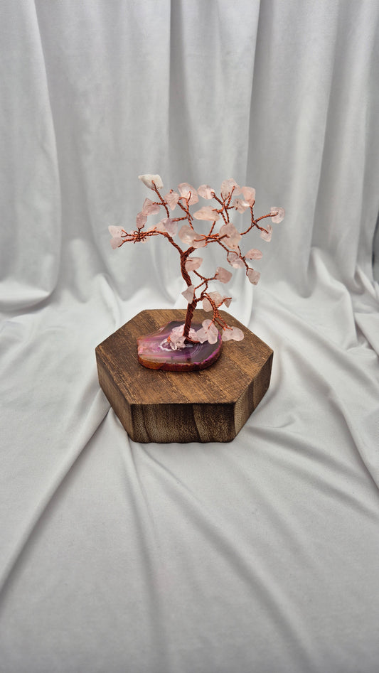 A cherry blossom crystal tree miniature with pink rose quartz blossoms and copper-colored branches, displayed on a polished purple agate slice and a hexagonal wooden block against a draped white fabric background.