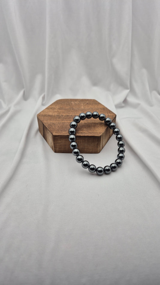 A beaded stretch bracelet made of glossy dark gray to black round pearls, draped over a hexagonal wooden block against a draped white fabric backdrop.