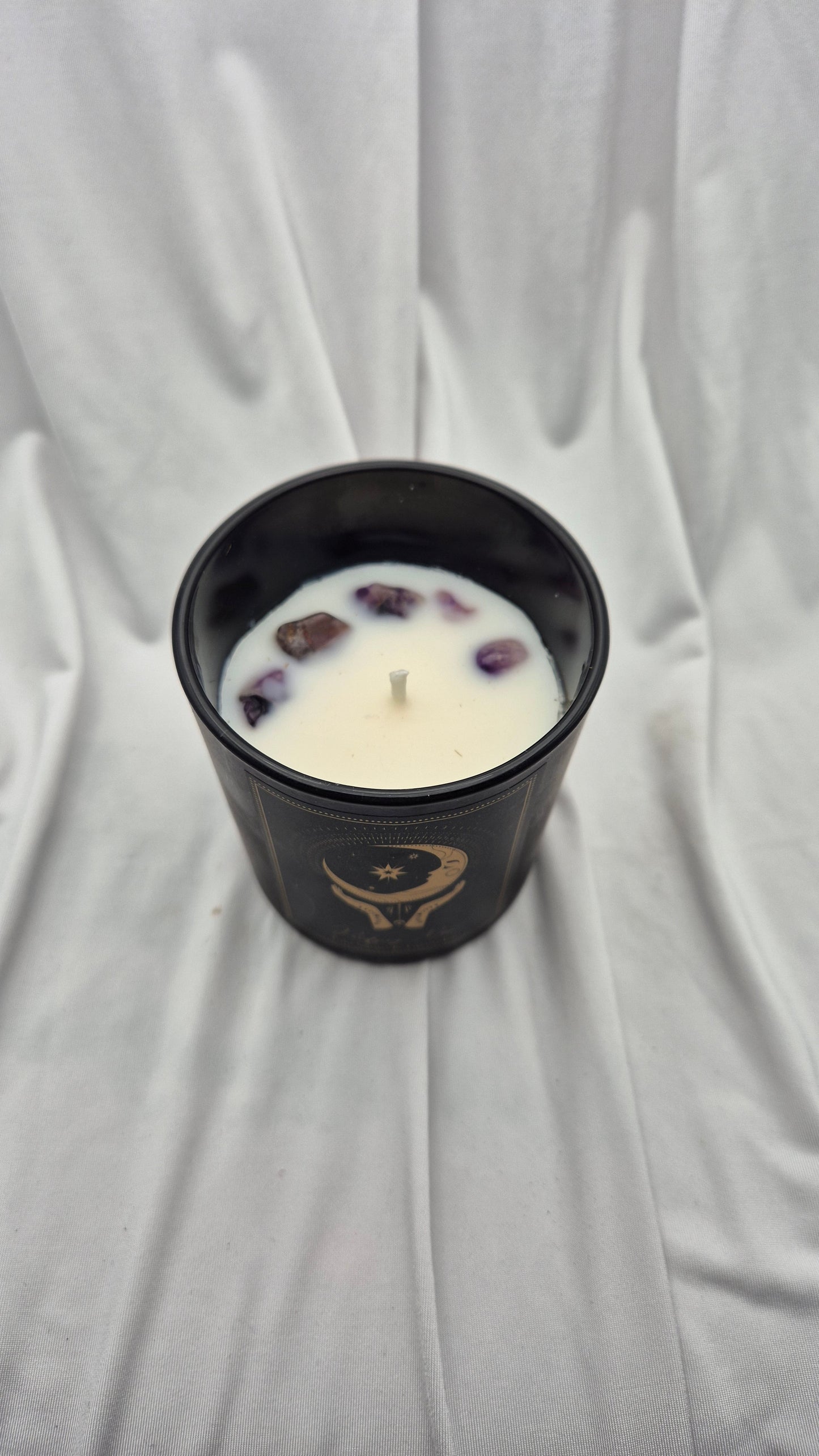 A black glass jar candle with a single wick, filled with white wax and decorated with small purple crystals. The jar features a gold crescent moon and star emblem on the front.