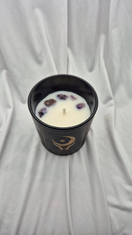 A black glass jar candle with a single wick, filled with white wax and decorated with small purple crystals. The jar features a gold crescent moon and star emblem on the front.