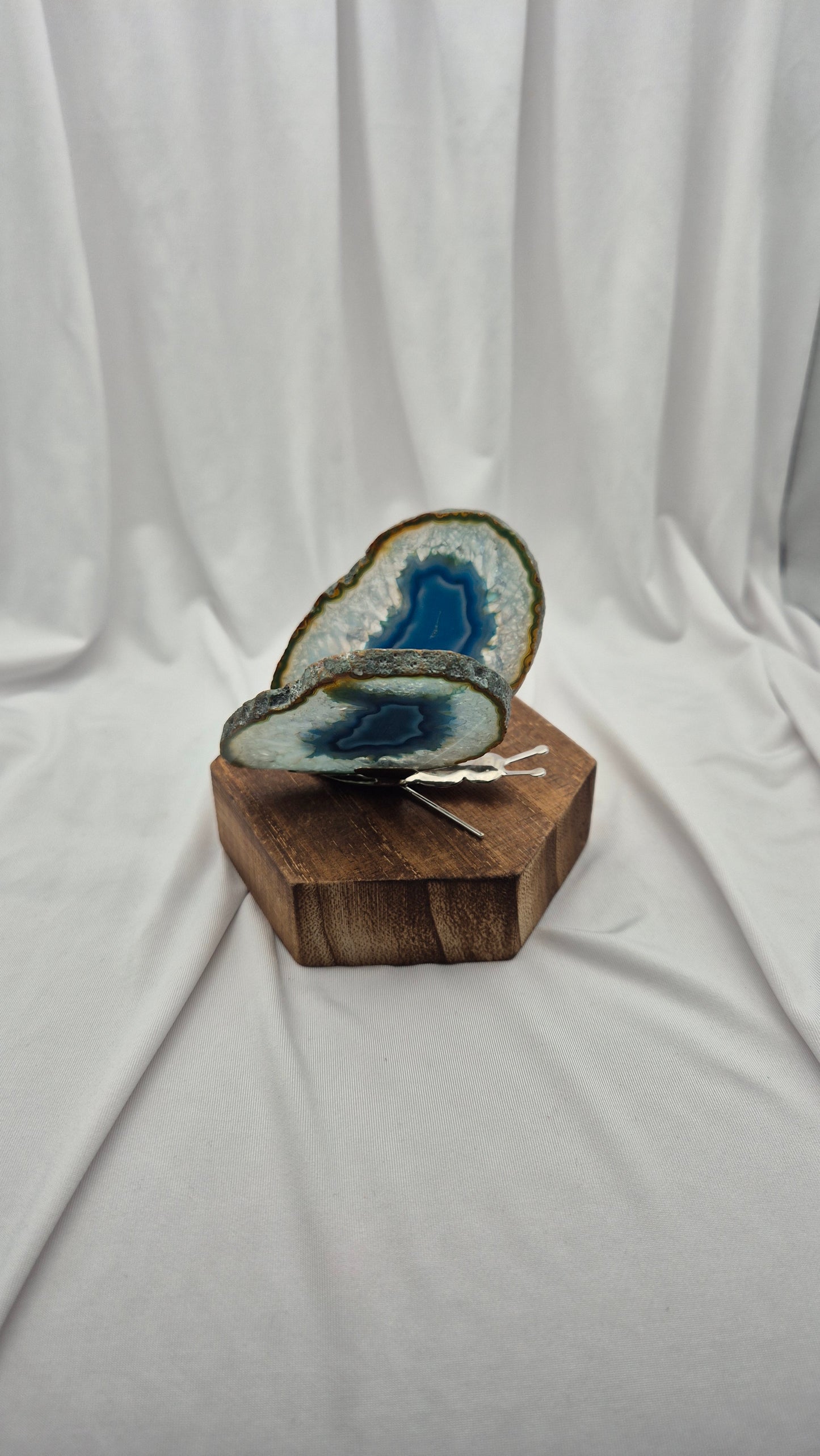 A polished blue agate geode slice with natural brown and white matrix, displayed on a hexagonal wooden block against a draped white fabric backdrop.