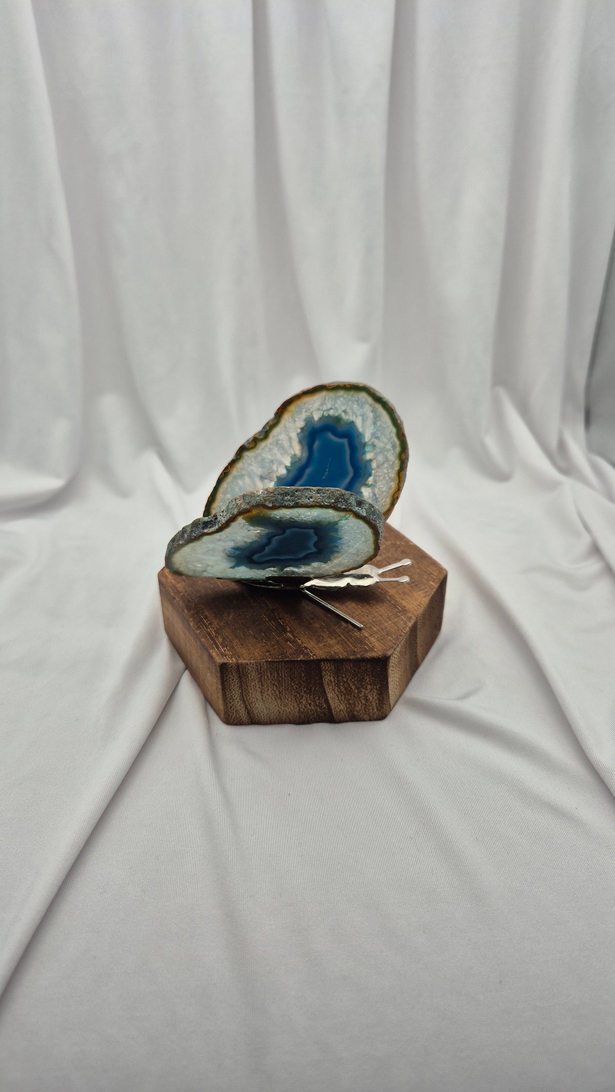 A polished blue agate geode slice with natural brown and white matrix, displayed on a hexagonal wooden block against a draped white fabric backdrop.