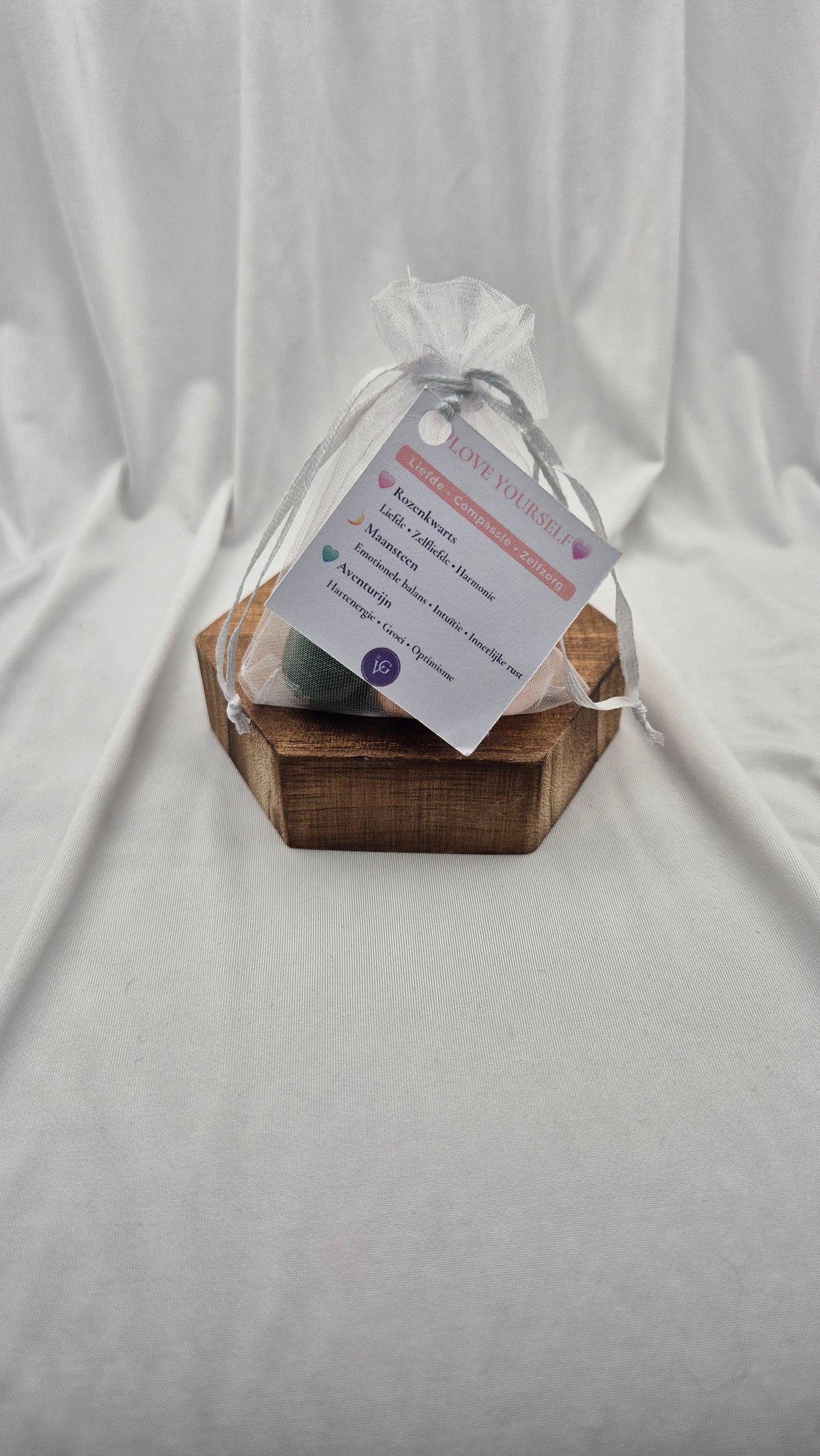 A small wooden hexagonal box holding a wrapped gift tied with a sheer white bag and a printed tag, set against a draped white fabric backdrop.