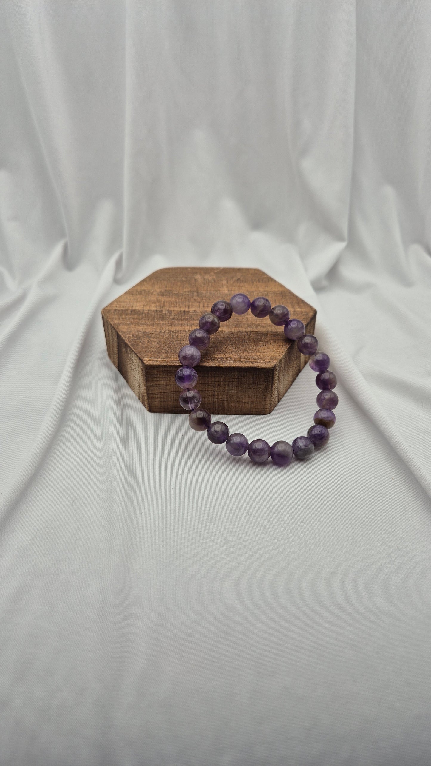 A purple beaded bracelet made of faceted round beads, displayed on a small hexagonal wooden block against a draped white fabric backdrop.