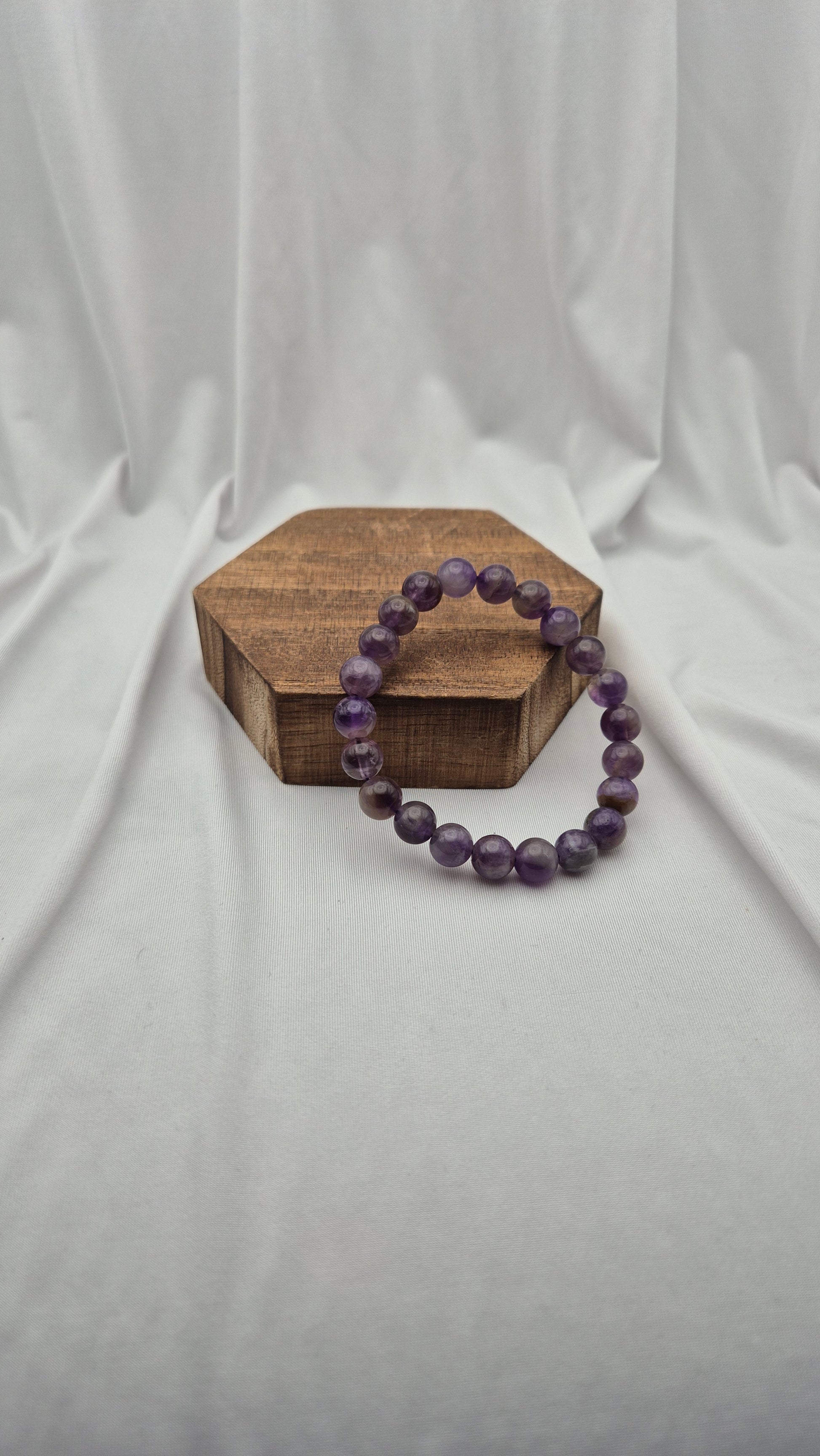 A purple beaded bracelet made of faceted round beads, displayed on a small hexagonal wooden block against a draped white fabric backdrop.