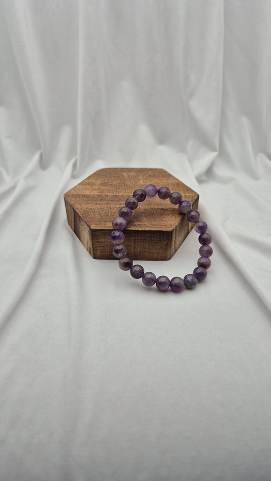 A purple beaded bracelet made of faceted round beads, displayed on a small hexagonal wooden block against a draped white fabric backdrop.