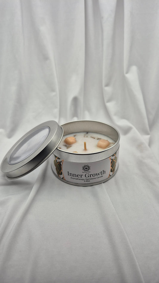 An open round metal tin candle labeled 'Inner Growth' on draped white fabric. The candle has white wax with two small peach-colored botanical pieces floating in it, and a silver lid leaning beside the container.