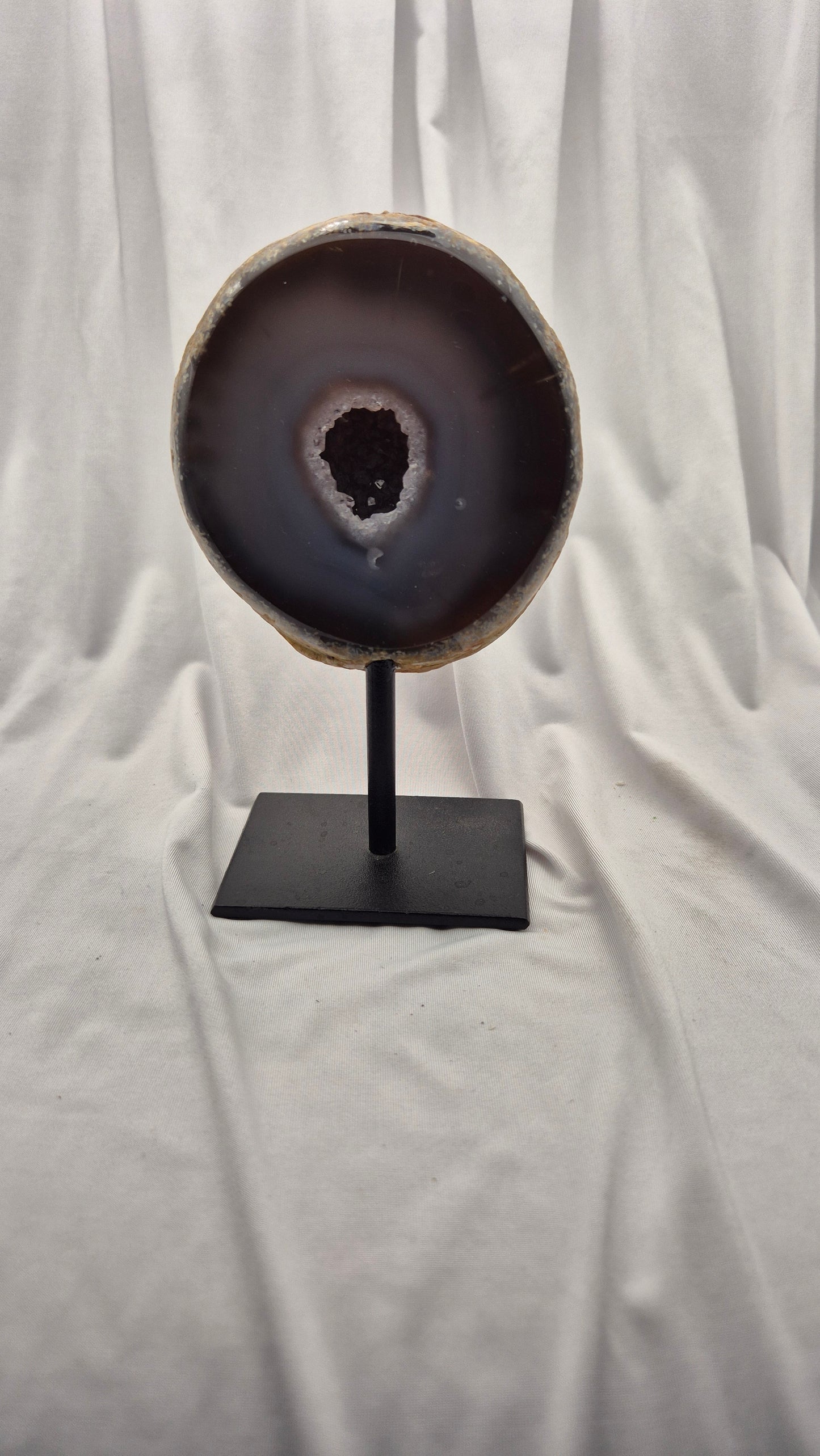 A polished agate geode slice with a dark brown and black outer band and a lighter beige inner rim, mounted on a black metal stand against a draped white fabric backdrop.