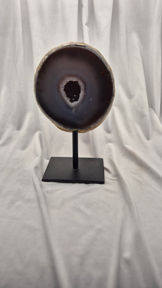 A polished agate geode slice with a dark brown and black outer band and a lighter beige inner rim, mounted on a black metal stand against a draped white fabric backdrop.